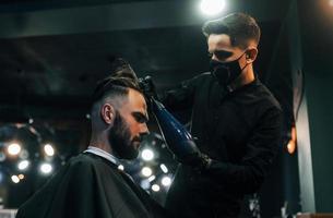 jovem barbudo sentado e cortando o cabelo na barbearia por cara com máscara protetora preta foto