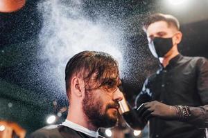 jovem barbudo sentado e cortando o cabelo na barbearia por cara com máscara protetora preta foto