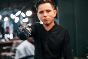 retrato de jovem em roupas pretas que está dentro de casa na barbearia e segurando uma tesoura foto