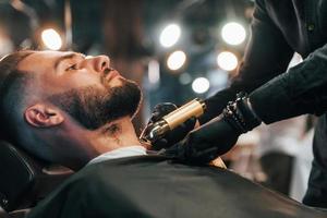 jovem com penteado estiloso sentado e raspando a barba na barbearia foto