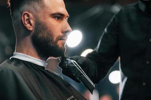 jovem com penteado estiloso sentado e raspando a barba na barbearia foto