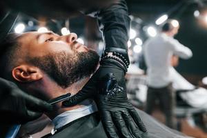 jovem com penteado estiloso sentado e raspando a barba na barbearia foto