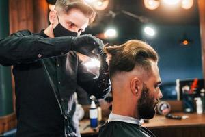 jovem barbudo sentado e cortando o cabelo na barbearia por cara com máscara protetora preta foto