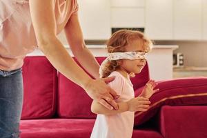 jogando jogo com os olhos vendados. jovem mãe com sua filha em roupas casuais juntos dentro de casa em casa foto