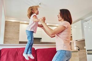 se divertindo e pulando. jovem mãe com sua filha em roupas casuais juntos dentro de casa em casa foto