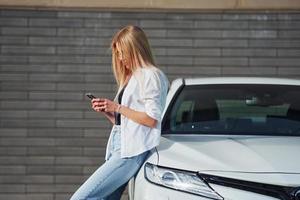 linda loira em roupas casuais com telefone perto de carro branco está ao ar livre durante o dia ensolarado foto