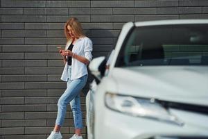 linda loira em roupas casuais com telefone perto de carro branco está ao ar livre durante o dia ensolarado foto