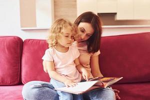 lendo livro grande. jovem mãe com sua filha em roupas casuais juntos dentro de casa em casa foto