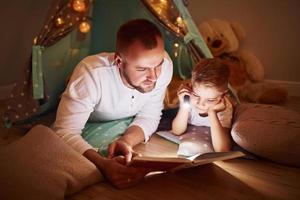 lendo livro e usando lanterna. pai e seus jovens passando tempo juntos dentro de casa perto da tenda foto