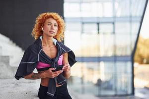 jovem ruiva europeia em roupas esportivas em pé na escada ao ar livre com o telefone nas mãos foto