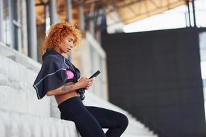 jovem ruiva europeia em roupas esportivas em pé na escada ao ar livre com o telefone nas mãos foto