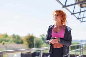 jovem ruiva europeia em roupas esportivas em pé ao ar livre com o telefone nas mãos foto