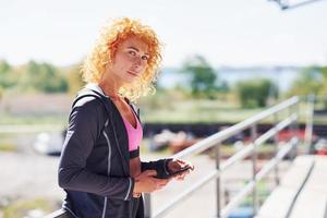 jovem ruiva europeia em roupas esportivas em pé ao ar livre com o telefone nas mãos foto