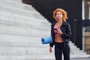 jovem ruiva europeia em roupas esportivas com telefone e tapete nas mãos ao ar livre foto