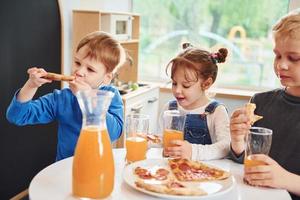 três crianças sentadas dentro de casa à mesa e comendo pizza com suco de laranja juntos foto
