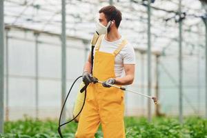 jovem trabalhador de estufa em uniforme amarelo e máscara protetora branca regando plantas usando equipamento especial dentro da estufa foto