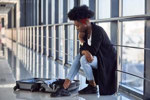 triste e deprimida jovem passageira afro-americana em roupas casuais, sentada no aeroporto com bagagem foto