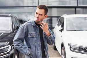 homem elegante fumando perto de carro ao ar livre contra edifício empresarial moderno foto