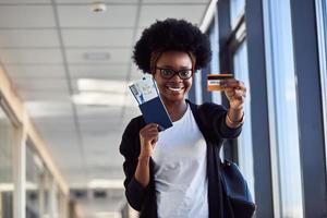 jovem passageira afro-americana em roupas casuais está no aeroporto segurando bilhetes e cartão de crédito foto