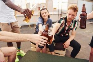 fazendo elogios por garrafas com cerveja. grupo de jovens em roupas casuais faz uma festa no telhado juntos durante o dia foto