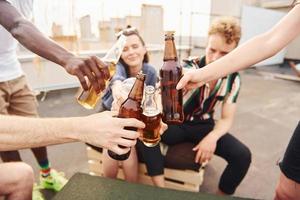 fazendo elogios por garrafas com cerveja. grupo de jovens em roupas casuais faz uma festa no telhado juntos durante o dia foto