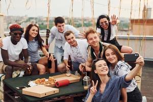 garota fazendo selfie. com deliciosas pizzas. grupo de jovens em roupas casuais faz uma festa no telhado juntos durante o dia foto