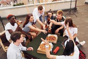 fazendo elogios por garrafas com cerveja. grupo de jovens em roupas casuais faz uma festa no telhado juntos durante o dia