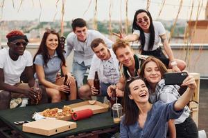 garota fazendo selfie. com deliciosas pizzas. grupo de jovens em roupas casuais faz uma festa no telhado juntos durante o dia foto