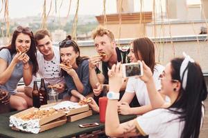 fazendo foto. deliciosa pizza. grupo de jovens em roupas casuais faz uma festa no telhado juntos durante o dia foto