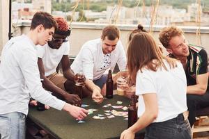 jogando e sentado à mesa. grupo de jovens em roupas casuais faz uma festa no telhado juntos durante o dia foto