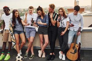 de pé com telefones e álcool nas mãos. grupo de jovens em roupas casuais faz uma festa no telhado juntos durante o dia foto