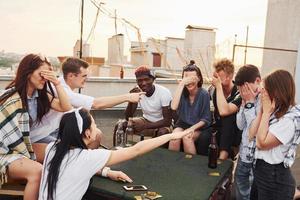 sentado à mesa e jogando jogo de cartas. grupo de jovens em roupas casuais faz uma festa no telhado juntos durante o dia foto