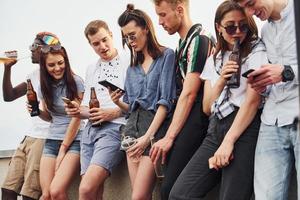 de pé com telefones e álcool nas mãos. grupo de jovens em roupas casuais faz uma festa no telhado juntos durante o dia foto