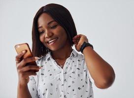 mulher afro-americana positiva na camisa branca em pé dentro de casa contra a parede branca com o telefone na mão foto