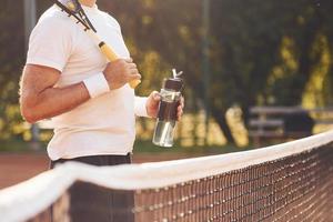 fazendo pausa e bebendo água. homem estiloso sênior em camisa branca e calção esportivo preto na quadra de tênis foto