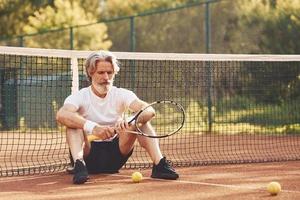 sentado e fazendo uma pausa. homem estiloso sênior em camisa branca e calção esportivo preto na quadra de tênis foto