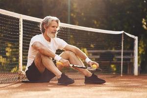 sentado e fazendo uma pausa. homem estiloso sênior em camisa branca e calção esportivo preto na quadra de tênis foto