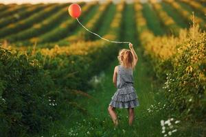 menina positiva com balão vermelho nas mãos divirta-se no campo durante o dia de verão foto