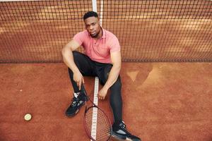 senta-se perto da rede e fazendo uma pausa. homem afro-americano de camisa rosa senta-se com raquete de tênis na quadra ao ar livre foto