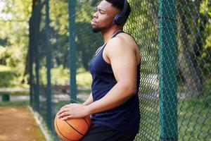 homem afro-americano com fones de ouvido sem fio faz uma pausa e apoia-se na malha de metal com bola na quadra ao ar livre foto