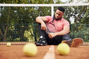 senta-se perto da rede e fazendo uma pausa. homem afro-americano de camisa rosa senta-se com raquete de tênis na quadra ao ar livre foto