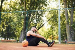 homem afro-americano cansado senta-se no chão com bola na quadra ao ar livre. faz uma pausa foto