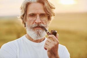 retrato de homem estiloso sênior com cabelos grisalhos e barba que está ao ar livre no campo em dia ensolarado e fumando foto