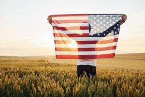 vista de trás. segurando a bandeira dos eua nas mãos. homem estiloso sênior patriótico com cabelos grisalhos e barba no campo agrícola foto