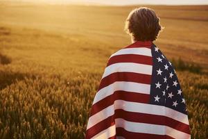 vista de trás. segurando a bandeira dos eua nas mãos. homem estiloso sênior patriótico com cabelos grisalhos e barba no campo agrícola foto