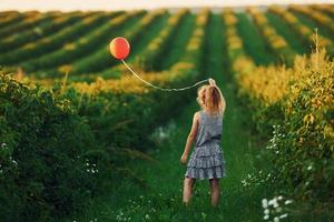 menina positiva com balão vermelho nas mãos divirta-se no campo durante o dia de verão foto