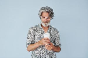 copo de bebida nas mãos. homem moderno elegante sênior com cabelos grisalhos e barba dentro de casa foto