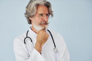 médico de jaleco branco contra a parede. homem moderno elegante sênior com cabelos grisalhos e barba dentro de casa foto