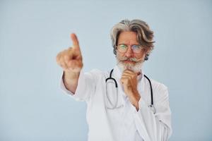 mostrando algo pelo dedo. trabalhador médico no casaco. homem moderno elegante sênior com cabelos grisalhos e barba dentro de casa foto