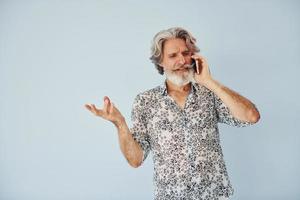 tendo chamada quando em pé contra a parede. homem moderno elegante sênior com cabelos grisalhos e barba dentro de casa foto